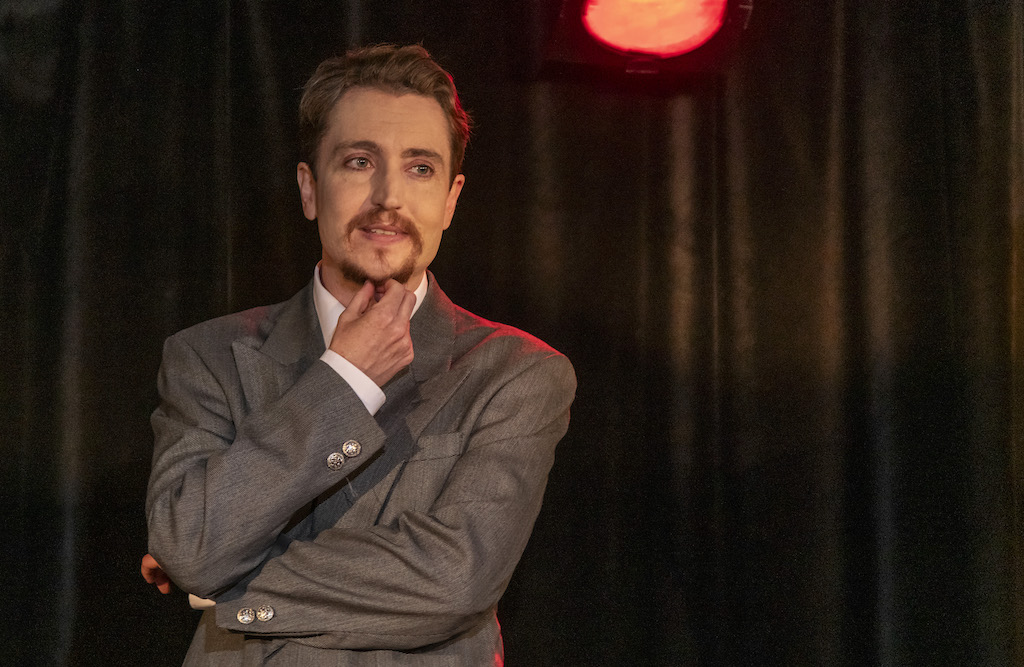 Head to waist photo showing Cloud Quinn onstage wearing a vintage grey suit. His arms are crossed and he strokes his goatee.