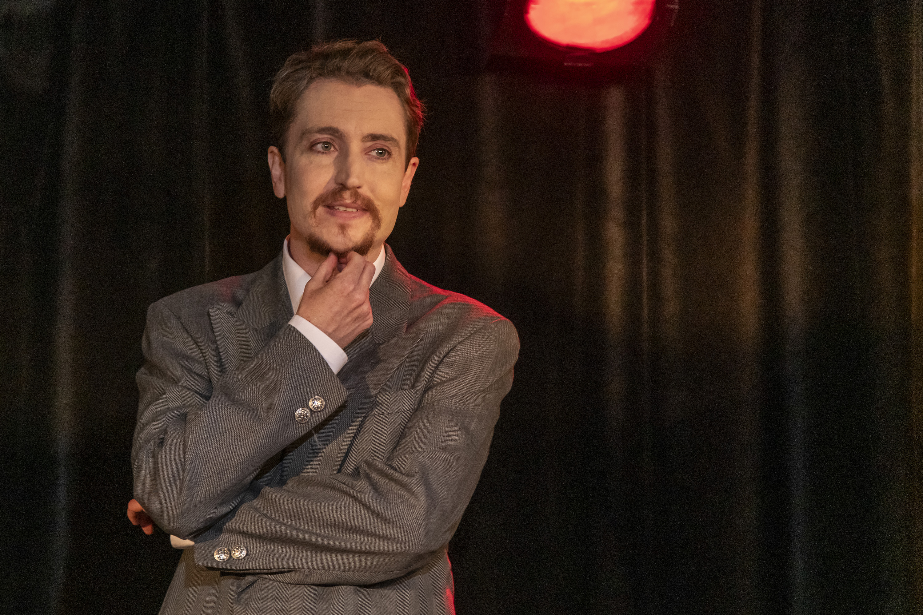 Head to waist photo showing Cloud Quinn onstage wearing a vintage grey suit. His arms are crossed and he strokes his goatee.