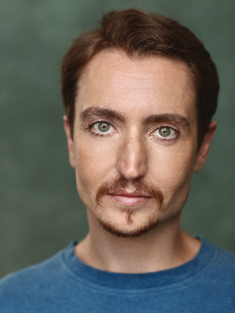 Head and shoulder photo of Cloud Quinn looking straight into the camera with a neutral expression. He has short brown hair and goatee and green-blue eyes. He wears an ocean blue T-shirt and stands in front of a teal background in a photographer’s studio.