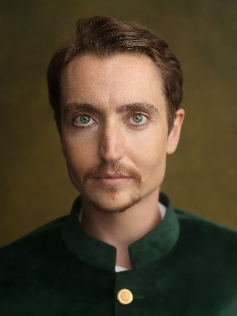 Head and shoulders photo of Cloud Quinn looking straight into the camera and smiling slightly. He has short brown hair and goatee and green-blue eyes. He wears a green velvet Nehru jacket with gold buttons and stands in front of a golden brown background in a photographer’s studio.
