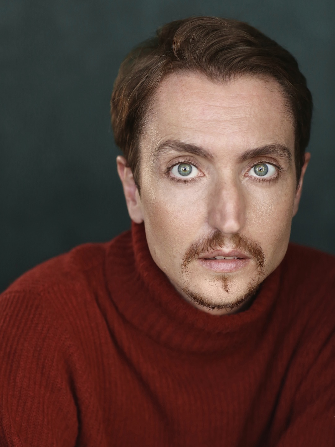 Head and shoulders photo of Cloud Quinn looking straight into the camera with a thoughtful expression, mouth slightly open. He has short brown hair and goatee and green-blue eyes. He wears a brick-red wool turtleneck jumper and stands in front of a slate blue background in a photographer’s studio.