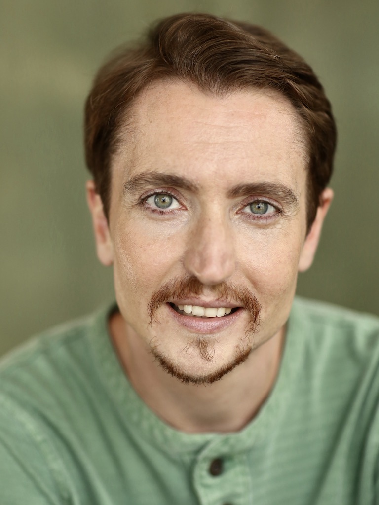 Head and shoulders photo of Cloud Quinn looking straight into the camera with a big smile, showing top row of teeth. He has short brown hair and goatee and green-blue eyes. He wears a sage green grandad collar long-sleeved T-shirt and stands in front of a forest green background in a photographer’s studio.