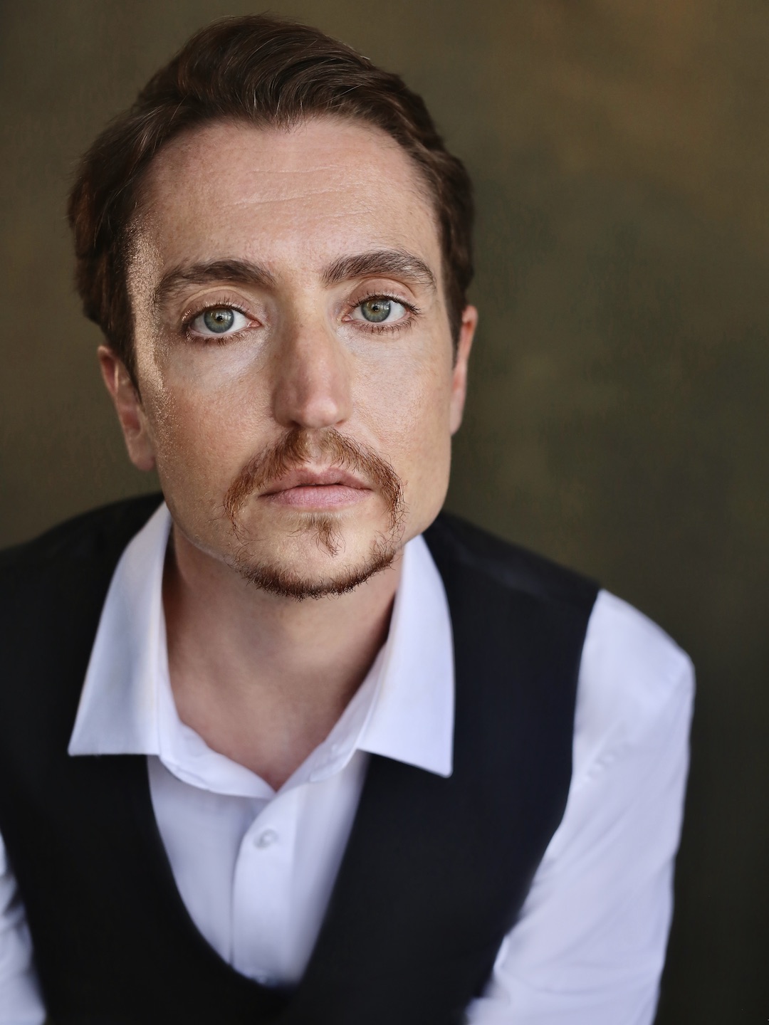 Head to mid-abdomen photo of Cloud Quinn looking straight into the camera with a distant expression. He has short brown hair and goatee and green-blue eyes. He wears a black waistcoat over a white button up shirt with collar and stands in front of a golden brown background in a photographer’s studio.
